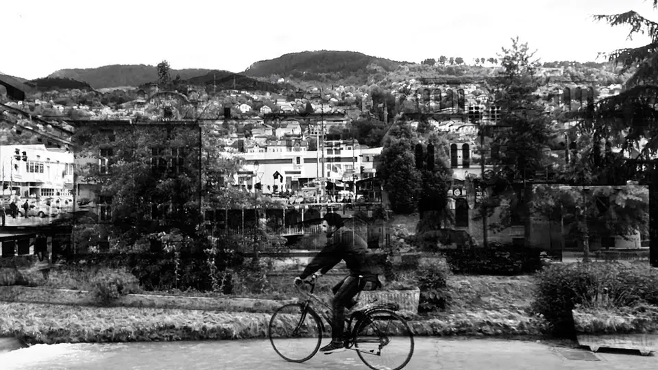 A double exposure in black and white: a man cycles past a riverside garden, the hillside houses of Sarajevo layered over the street