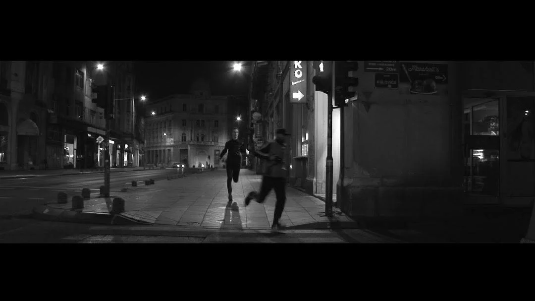 Photostory: two men run down an empty street in Sarajevo at night, in black and white, between the black bars of a widescreen frame