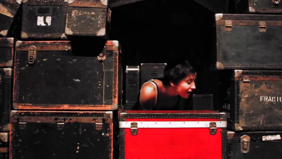 A performer leans out from a wall of stacked old travel trunks, a red trunk in front of her