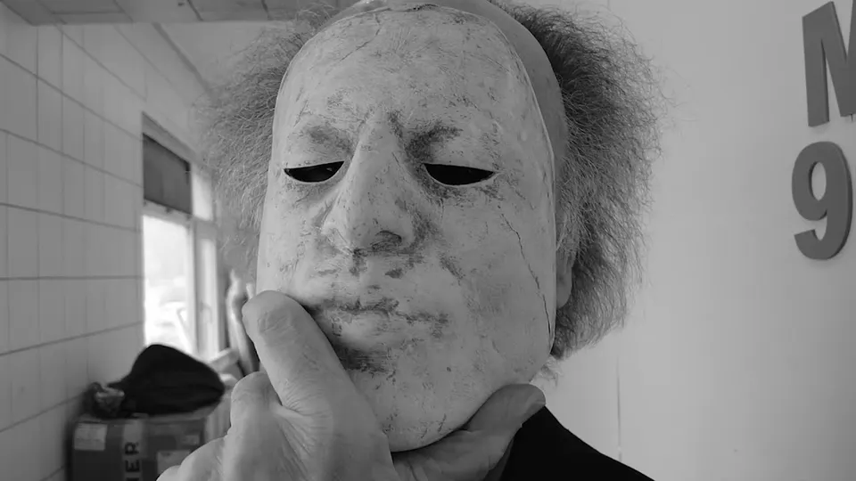 A hand holds a pale, cracked mask of an old man's face to the chin, in a white-tiled room