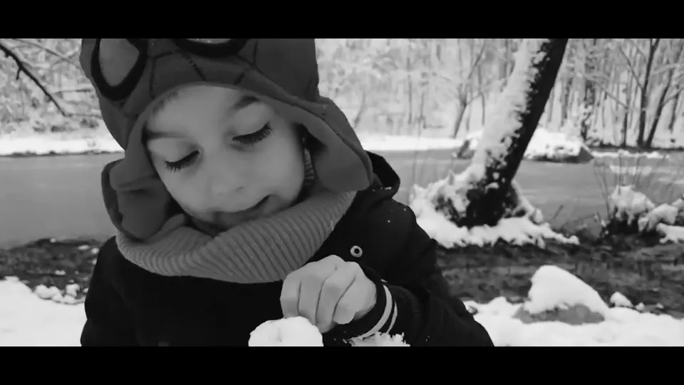 A small boy in a winter hat looks down at a snowball in his hands, a snowy river behind him, in black and white